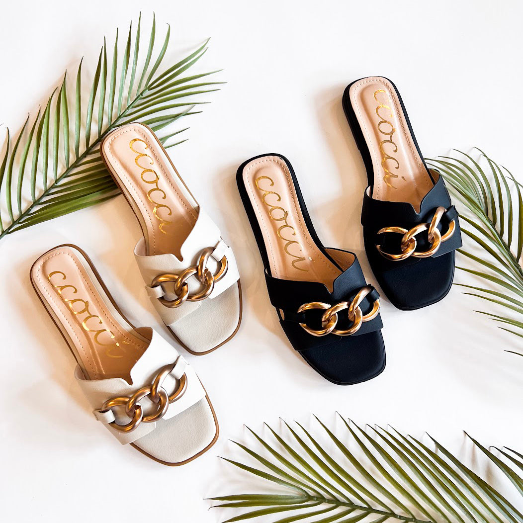 Two pairs of Baylor sandals with chunky gold chain accents, one in white and the other in black, arranged on a white background with green palm leaves for a stylish, tropical aesthetic