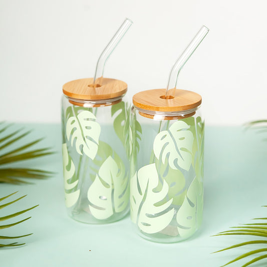 Eco-friendly glass tumbler featuring a green monstera leaf design, complete with a bamboo lid and glass straw, set against a light background with tropical palm leaves