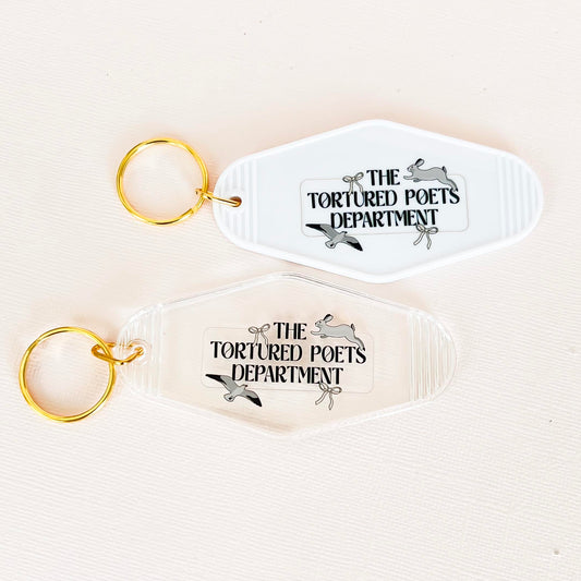 "Two vintage-inspired keychains labeled 'The Tortured Poets Department.' One is white with a golden keyring, and the other is clear with a golden keyring. Both feature a unique, playful design perfect for literary enthusiasts."