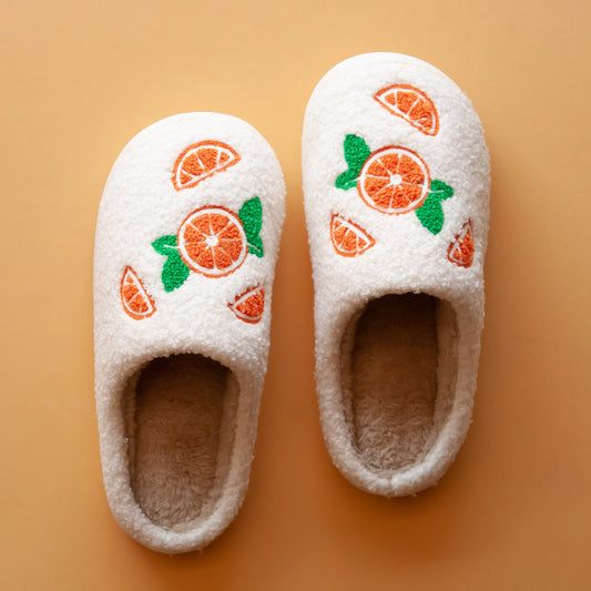 White cozy slippers with fuzzy texture, adorned with embroidered orange slices and green leaves on the top. 